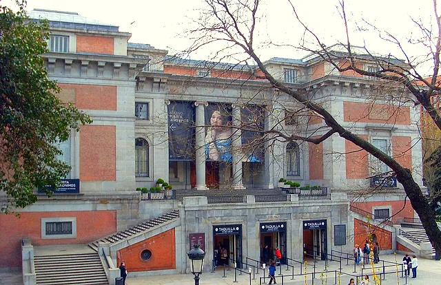 A side entrance of the Prado Museum, with red brick and stone walls, a large banner and visitors at the doors