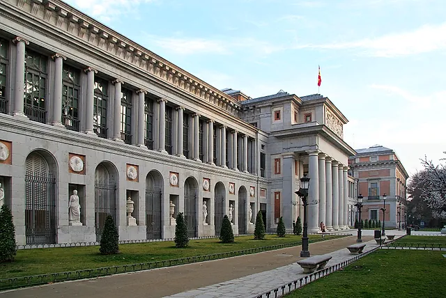 The long façade of the Prado Museum along its garden path, under a pale sky